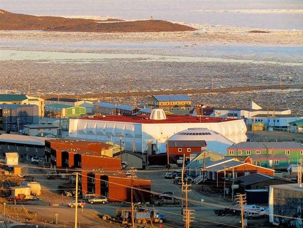 A small coastal town in Nunavut featuring a mix of industrial, residential, and public buildings with ice-covered waters and distant shore in the background.