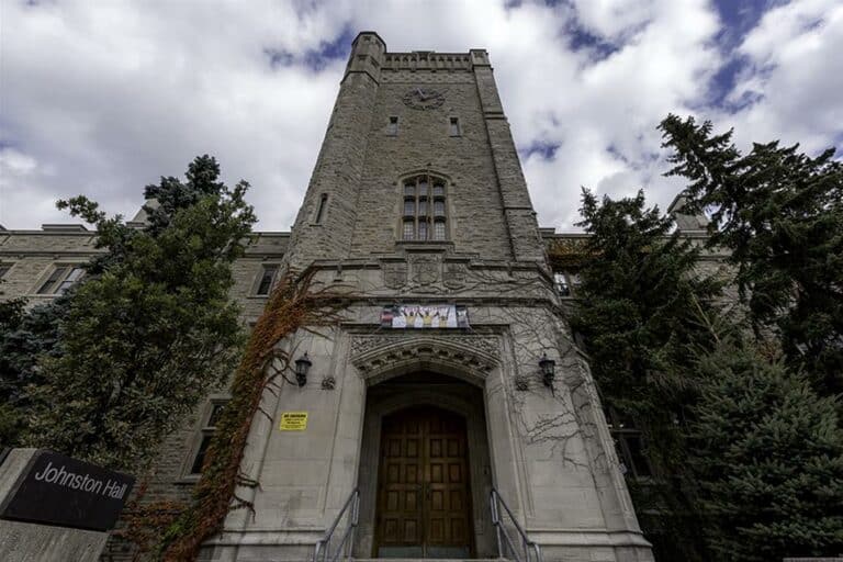 A tall, stone building with a prominent, arched entrance and an inscription above the door. The sign "Johnston Hall" is visible on the left, marking its stature within the University of Guelph. Trees surround the building under a partly cloudy sky, embodying the spirit of higher education.