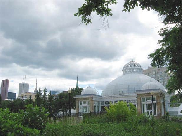 White conservatory with a glass dome set in a green garden, with tall buildings and a cloudy Toronto sky in the background.