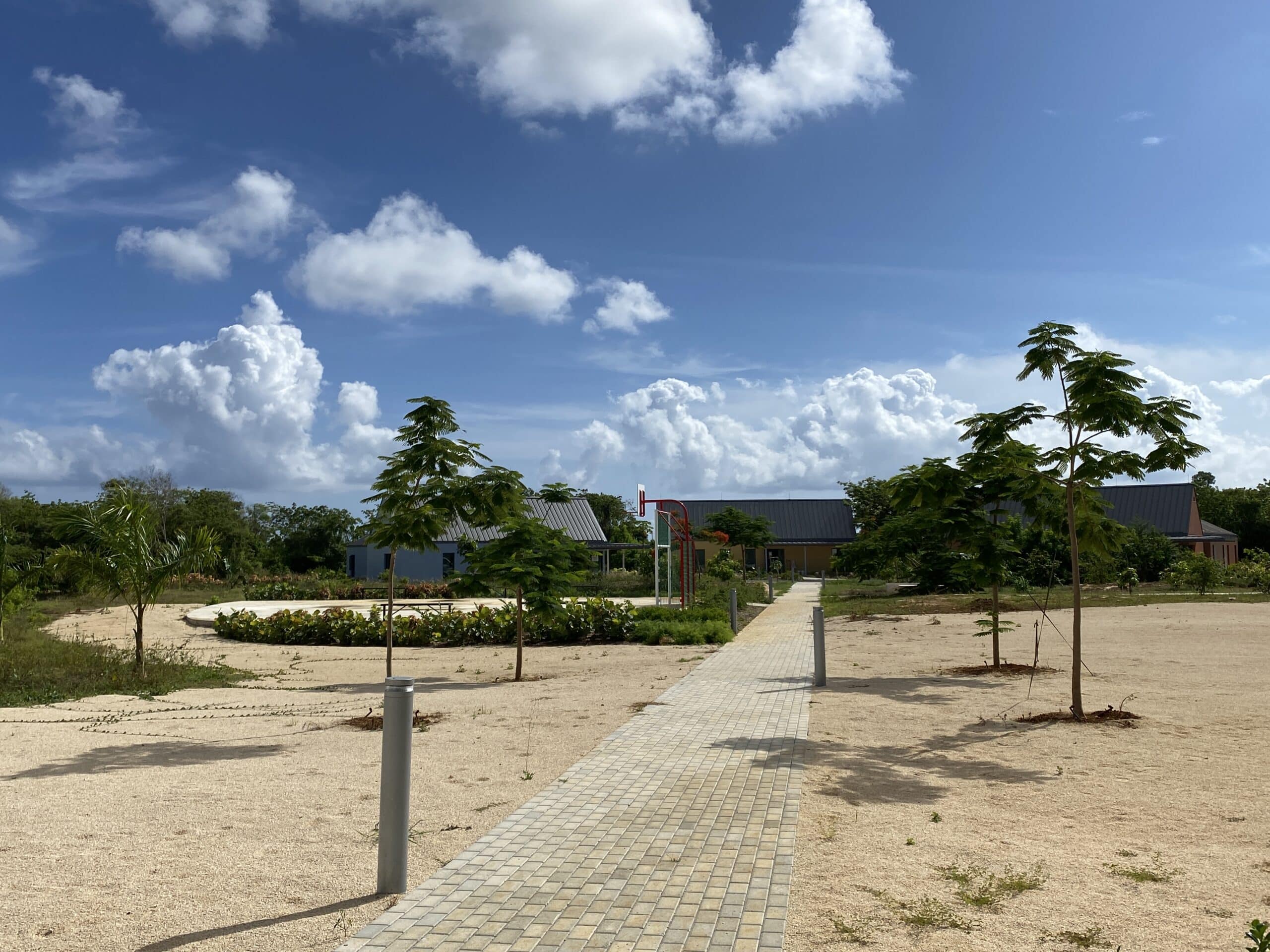Exterior view of the Poinciana Rehabilitation Centre, showing multiple buildings, trees, and a wide walkway surrounded by sand.
