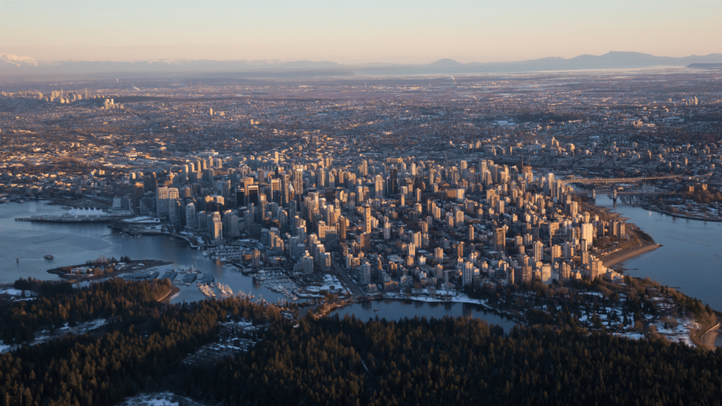Aerial view of downtown Vancouver at sunset, with high-rise buildings surrounded by water, forested parkland in the foreground, and mountains in the distance.