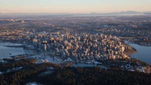 Aerial view of downtown Vancouver at sunset, with high-rise buildings surrounded by water, forested parkland in the foreground, and mountains in the distance.