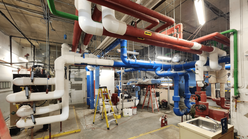 Commercial mechanical room with colour-coded HVAC piping system, including blue chilled water lines, red heating pipes, white insulated pipes, and green supply lines, during active retrofit construction in a Canadian commercial building.